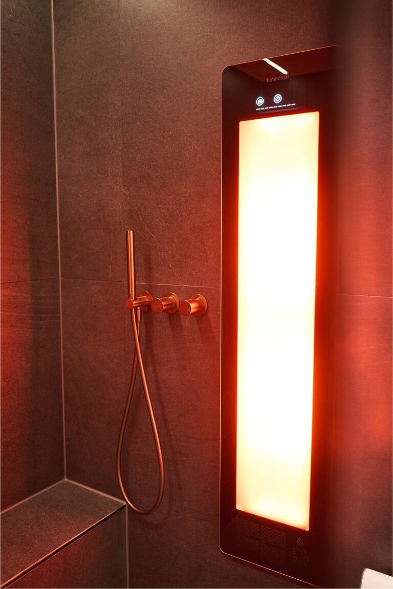Infrarotstrahler leuchtet rot-gelb und ist an der Duschwand, neben den Armaturen aus Bronze, angebracht. Die Fliesen im Hintergrund sind dunkelgrau.