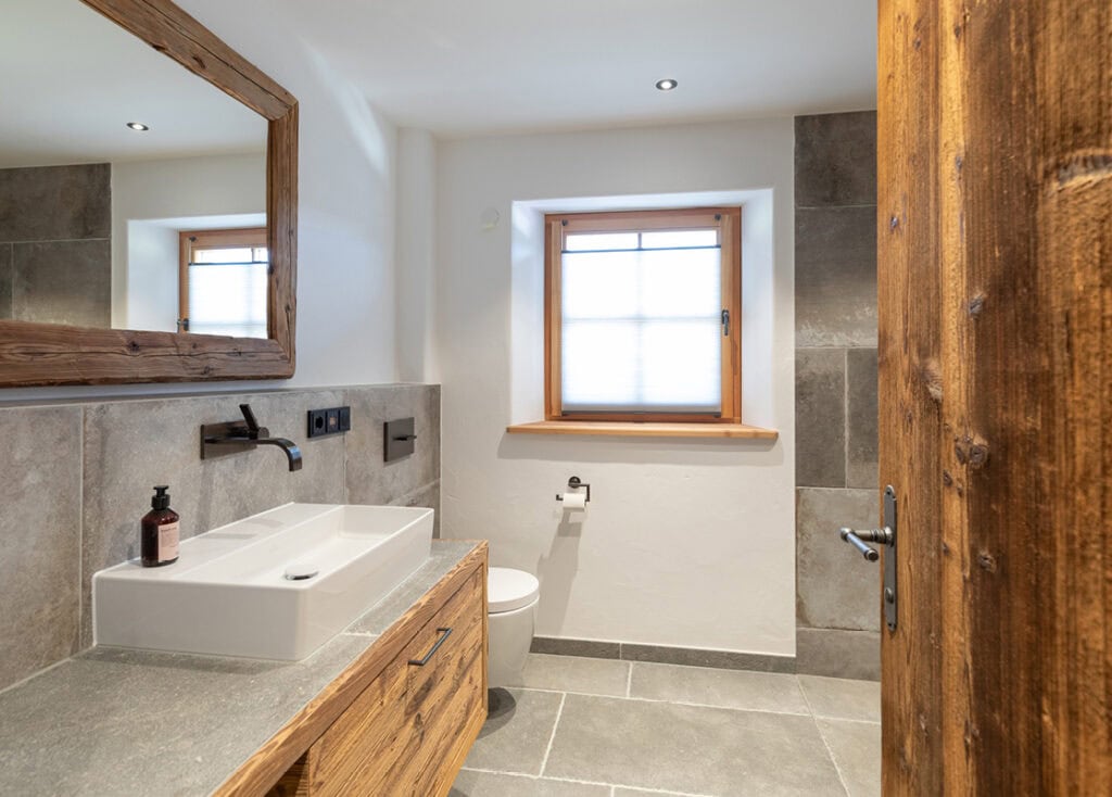 Modernes Bad mit hellgrauen Fliesen und Altholzelementen. Das weiße Waschbecken aus Keramik ist viereckig und wurde auf den Unterschrank aufgesetzt.