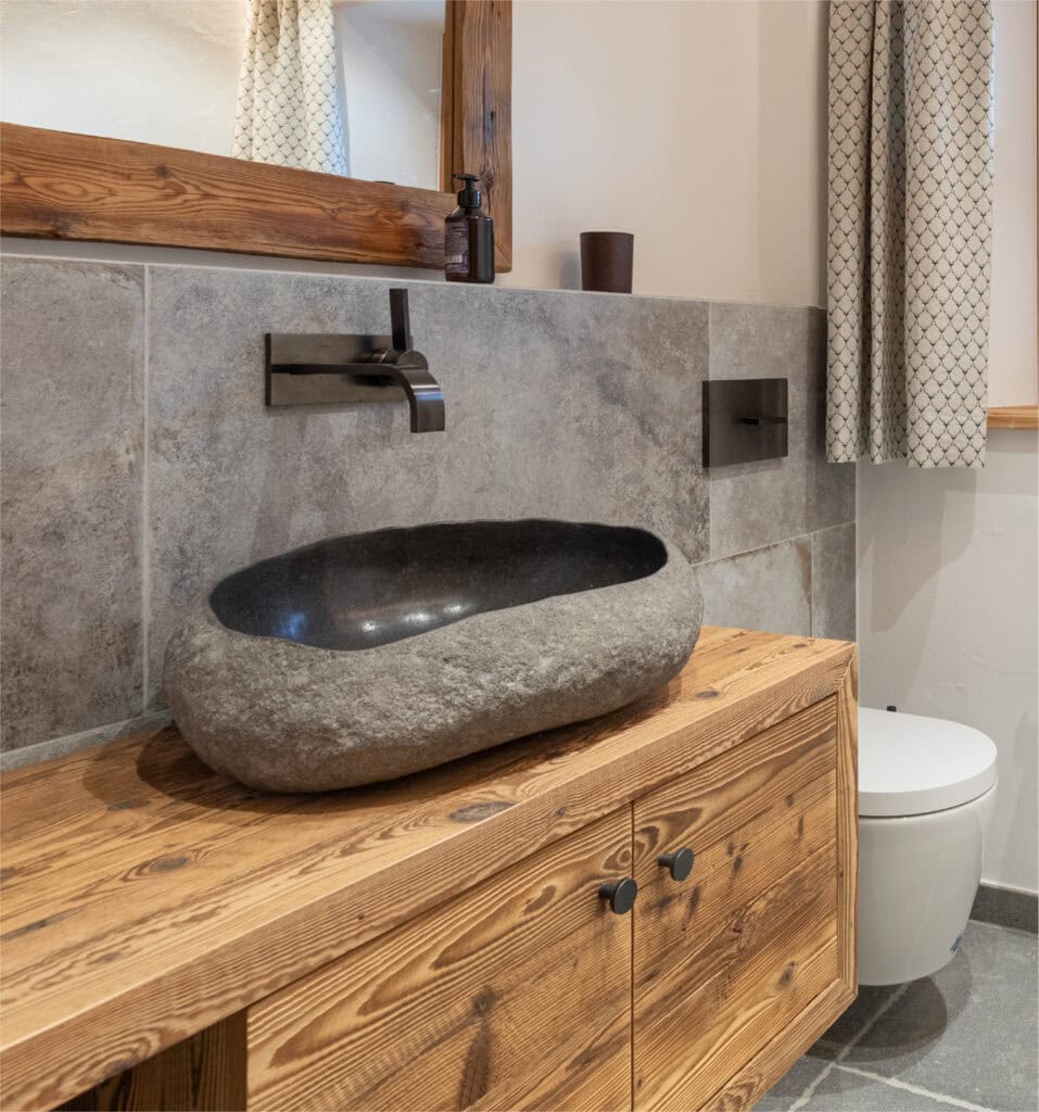 Ovaler, graues, aufgesetztes Steinwaschbecken mit an der Wand angebrachten schwarzen, modernen Wasserhahn. Das Waschbecken steht auf einem Altholz-Unterschrank.