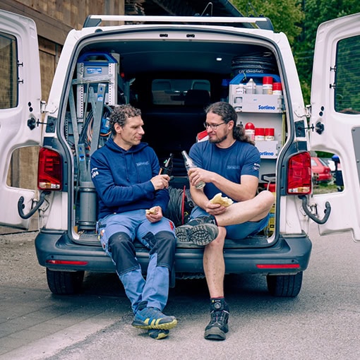 Zwei Männer sitzen an der Heckklappe des Autos und machen Brotzeit.