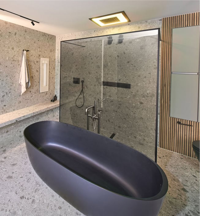 Modernes Bad mit Steinfliesen am Boden und an den Wänden mit einer freistehenden matt-schwarzen Badewanne. Im Hintergrund befindet sich eine ebenerdige Dusche mit einer Glastrennwand.