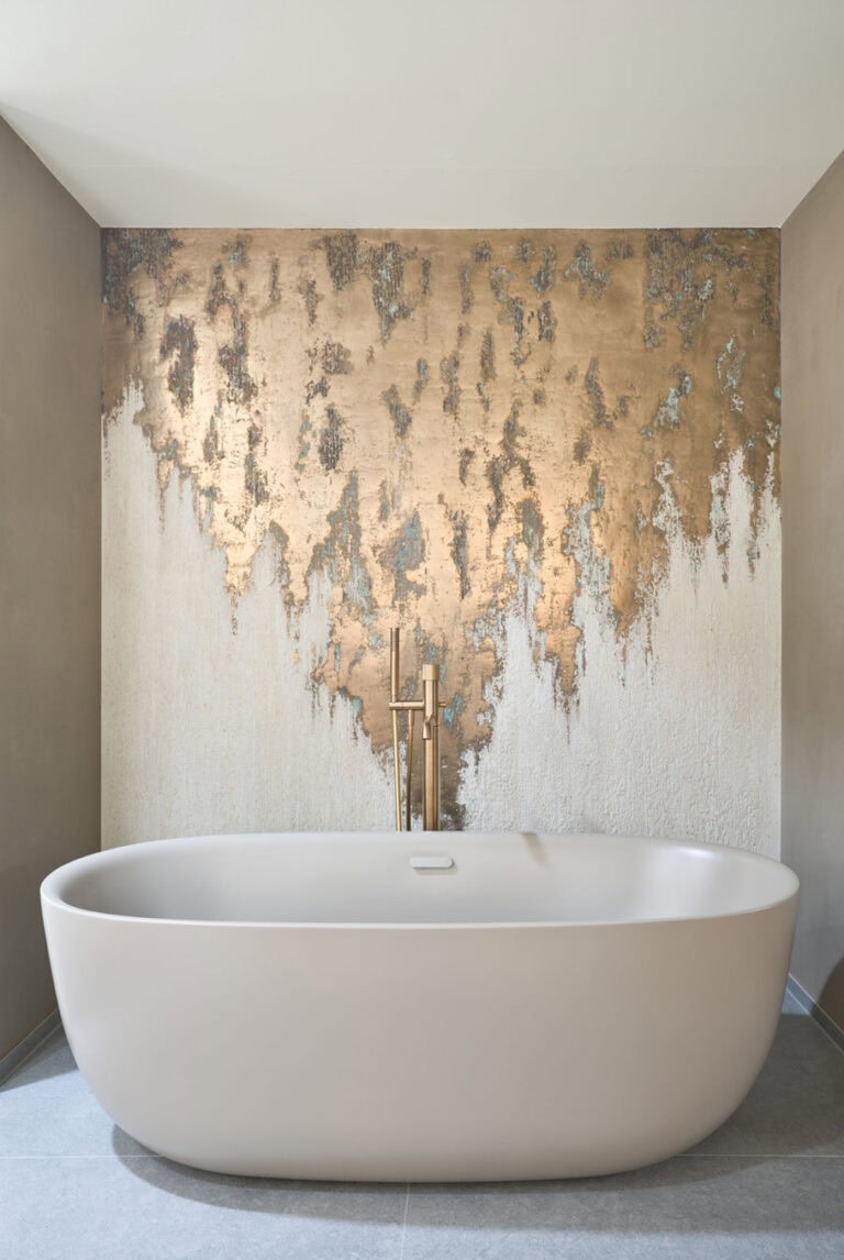 Cremefarbige, freistehende Badewanne mit goldener Wand im Hintergrund.