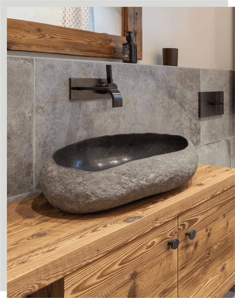 Ovaler, graues, aufgesetztes Steinwaschbecken mit an der Wand angebrachten schwarzen, modernen Wasserhahn. Das Waschbecken steht auf einem Altholz-Unterschrank.