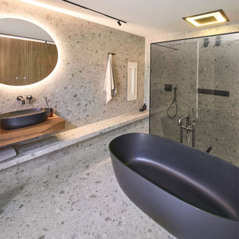 Luxusbad mit Steinboden und Steinwand, einem hochwertigen aufgesetztem, schwarzem Waschbecken auf einer Holzplatte sowie einer freistehenden schwarzen Badewanne und eine ebenerdige Dusche mit Infrarotstrahler.