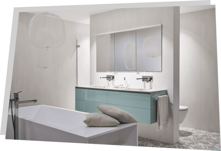 Ein neues Bad mit heller Fliesenoptik. Im Hintergrund befindet sich ein Waschbecken mit Spiegel und Hängeschrank sowie Waschbecken. Im Vordergrund eine Badewanne auf der zwei Kissen trapiert sind.